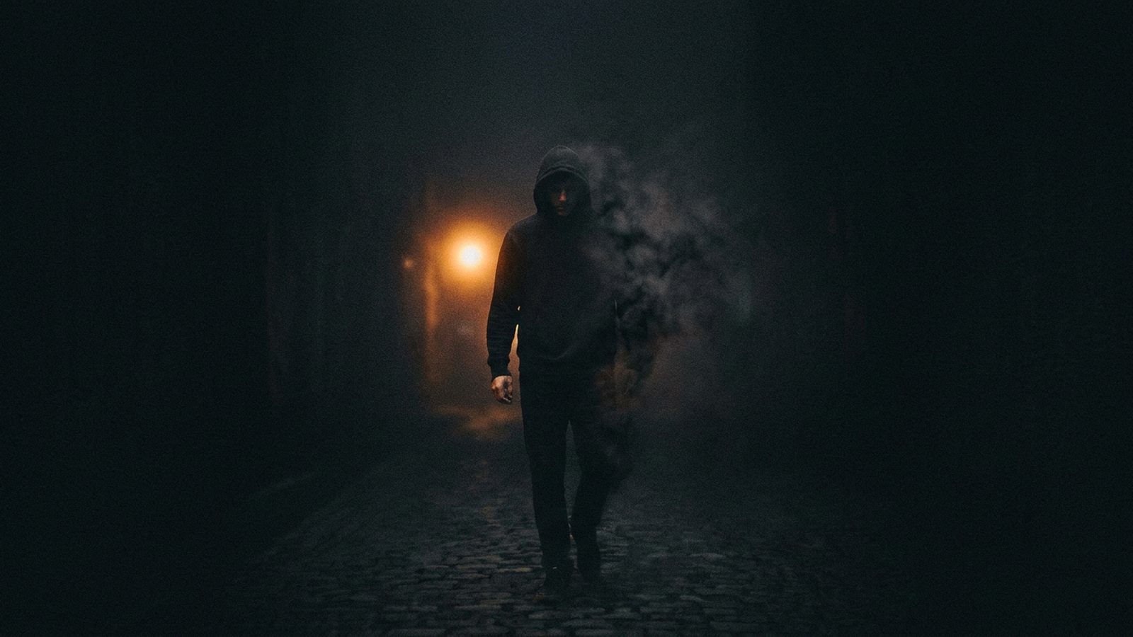 A hooded man walking alone down a dark, foggy street toward a single streetlight, retracing the same path again
